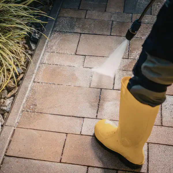 Child cleaning outdoor brick patio with a hose, wearing yellow rain boots.