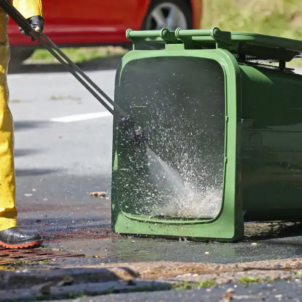 Green garbage can being pressure washed outdoors for cleaning.